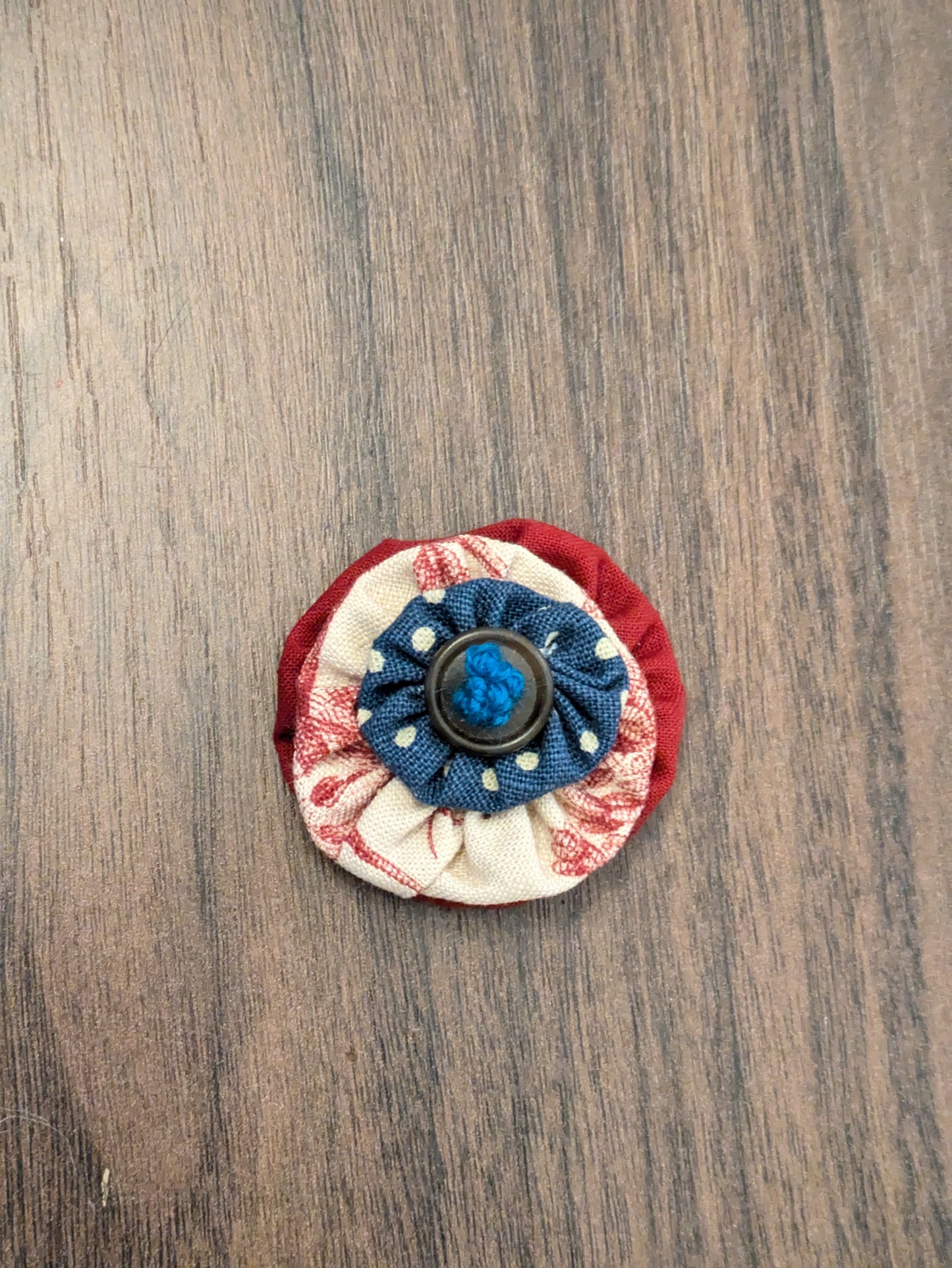 A material flower with a base red layer, a second layer of with cream with red flowers and a small blue layer with white spots, finished with a brown button and hand-embroidered centre with dark turquoise thread. Made to thread onto a dogs collar.