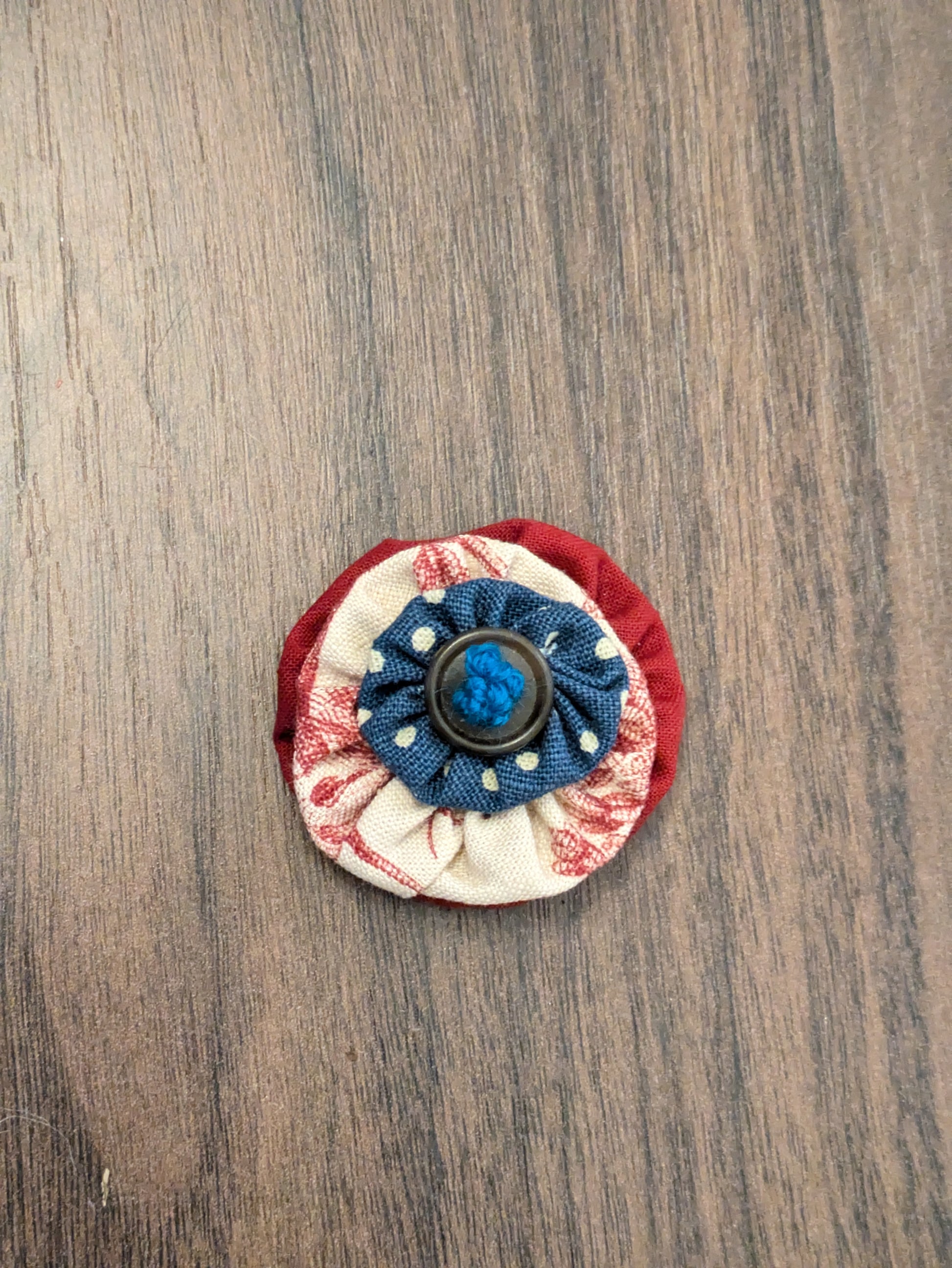 A material flower with a base red layer, a second layer of with cream with red flowers and a small blue layer with white spots, finished with a brown button and hand-embroidered centre with dark turquoise thread. Made to thread onto a dogs collar.